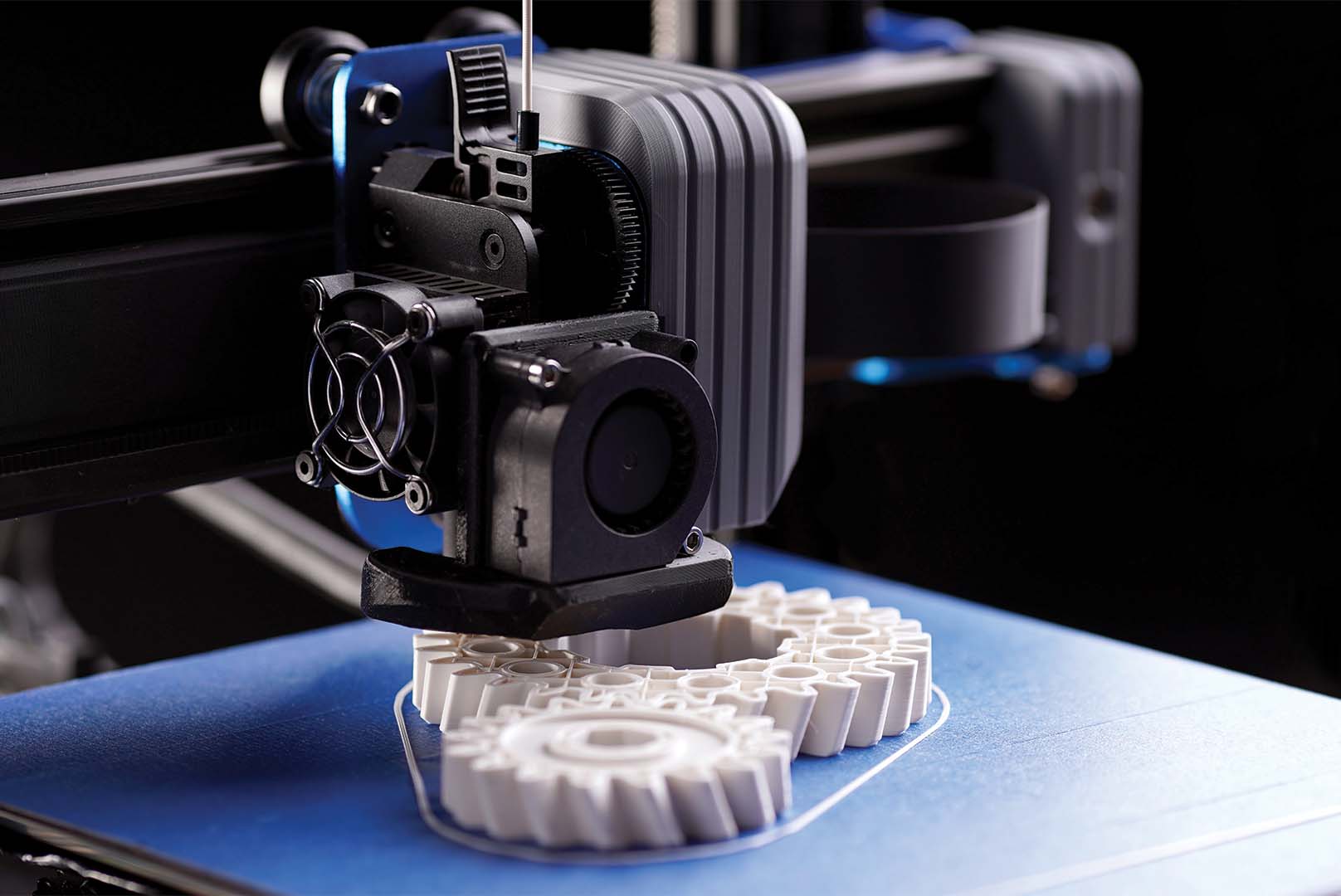 FDM 3D printer print head extruding white PLA gears onto a blue textured build plate